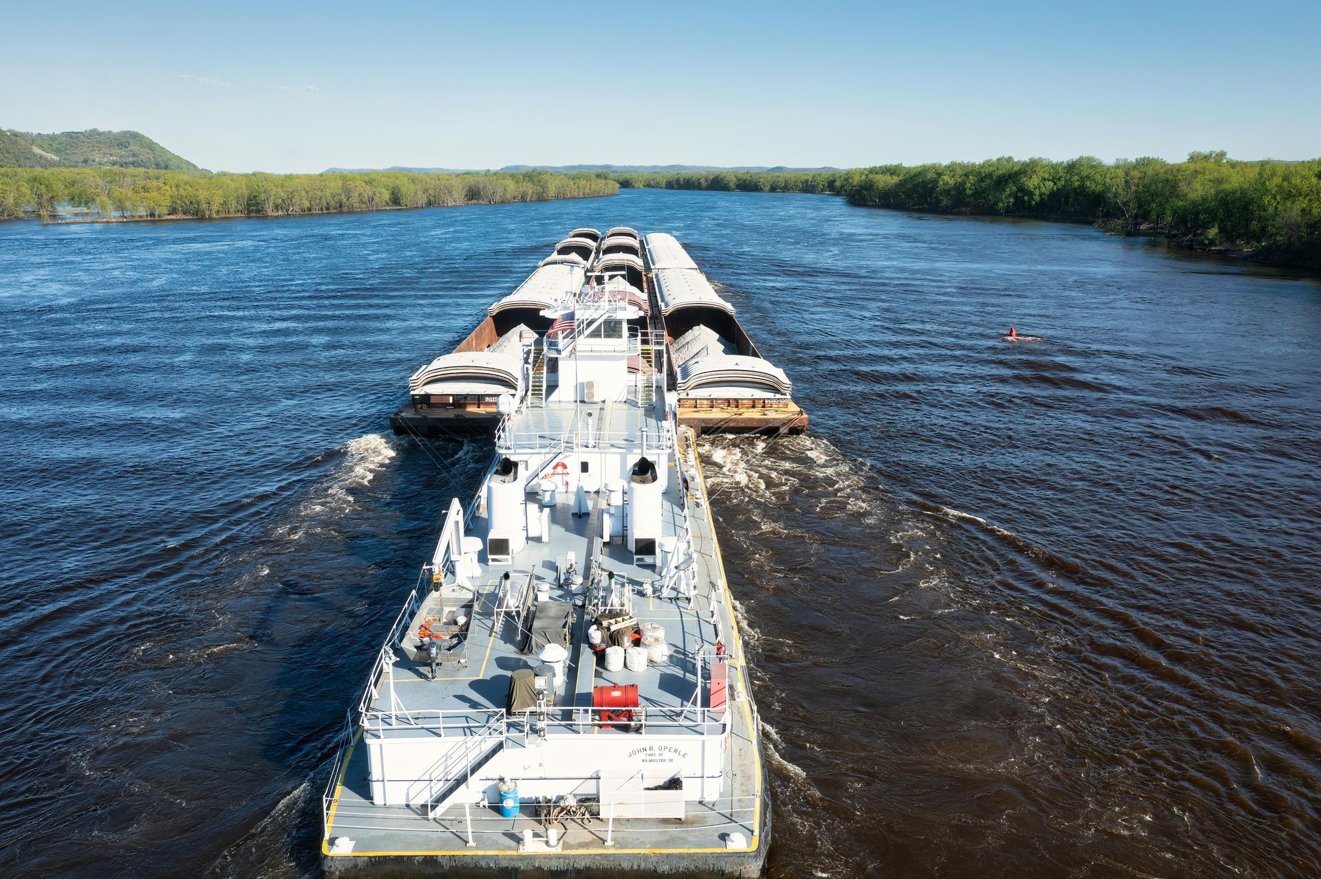 Tugboat pushing a long barge down a wide river under a clear blue sky