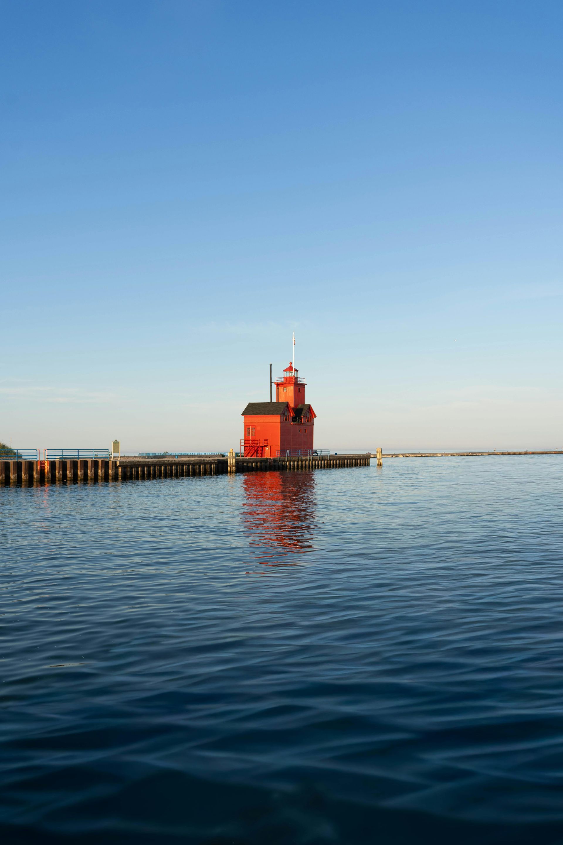 Red lighthouse on a calm blue sea at sunset, with a pier stretching into the distance