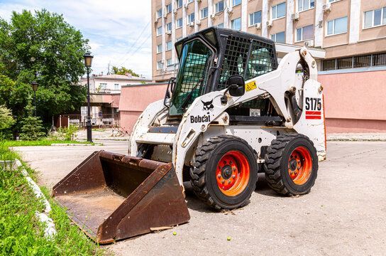 White Bobcat S175 skid-steer loader with rusty bucket; parked near building and trees.