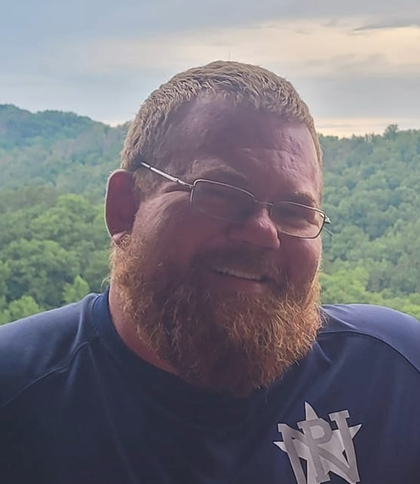 Man with a full beard and glasses smiling outside, mountains in the background.