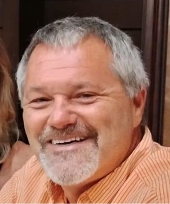 Man with graying hair and beard smiles, wearing an orange striped shirt.