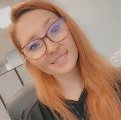Woman with red hair and glasses smiles at the camera, indoors.
