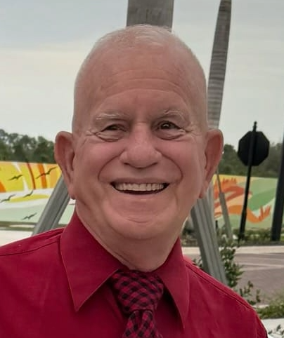 Smiling man in a red shirt and patterned tie stands outside, with a mural in the background.