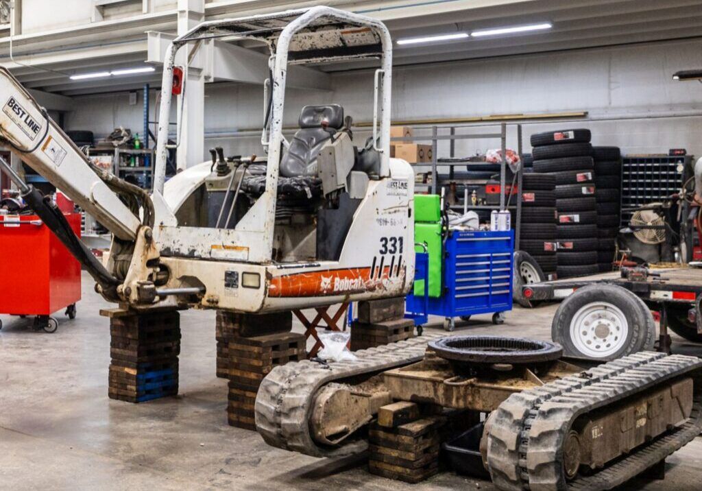 A Bobcat excavator being worked on inside a garage, track treads and tires visible.
