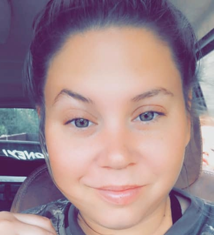 Woman with light eyes smiles, hair up, sitting in a vehicle.