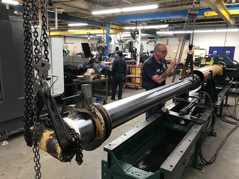 Man works on machinery in a shop; a large cylinder is suspended by chains.