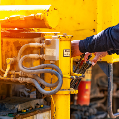 Close-up of a yellow construction machine. A person's hand operates a lever, likely for hydraulic controls.