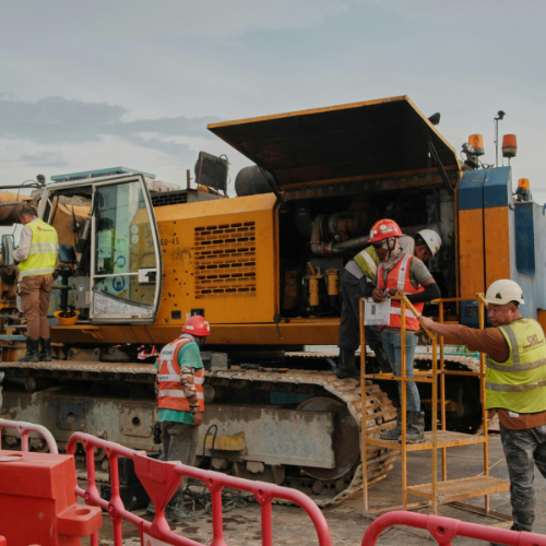 Construction workers inspecting a yellow and blue excavator on a worksite; wearing safety vests and helmets.