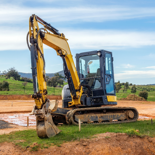 Yellow excavator on a construction site with a blue sky and green fields in the background.