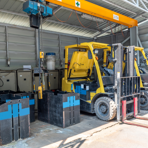 Yellow forklifts parked in a warehouse with batteries in blue containers under an overhead crane.