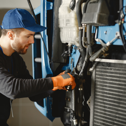 Mechanic in blue cap and orange gloves, working on a vehicle's engine.