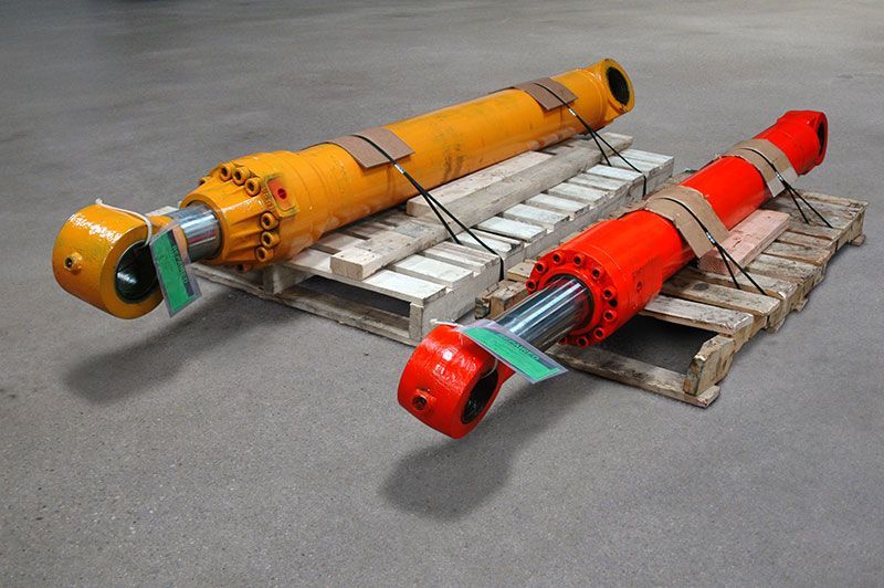 Two hydraulic cylinders, one yellow, one red, strapped to wooden pallets.