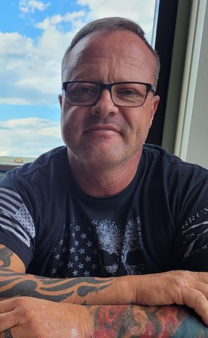 Man with glasses, tattoos, and black shirt, looking at the camera outdoors, with a sky background.