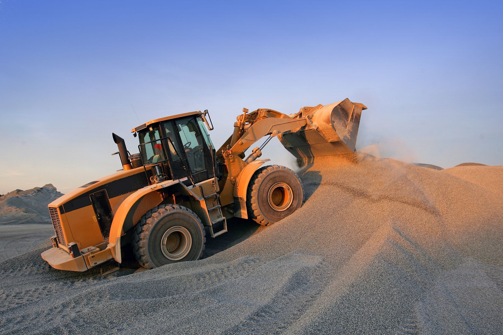 Yellow excavator on a construction site with a blue sky and green fields in the background.