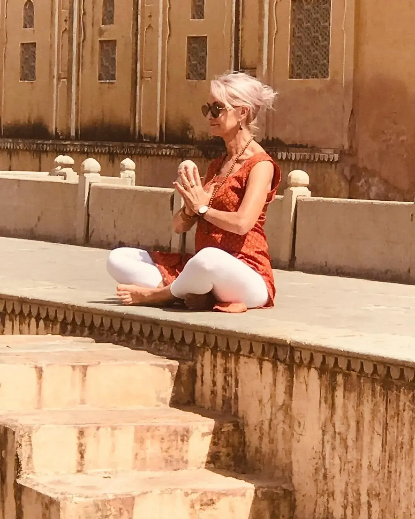 A person in a patterned tunic and white pants sits on a green railing with the Taj Mahal in the background at sunset.