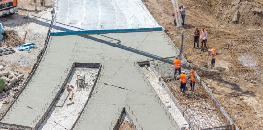 Construction workers pour concrete into a large, Y-shaped foundation form on a dirt work site.