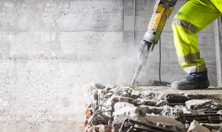 A construction worker in a high-visibility yellow uniform uses a jackhammer to break up a concrete floor.