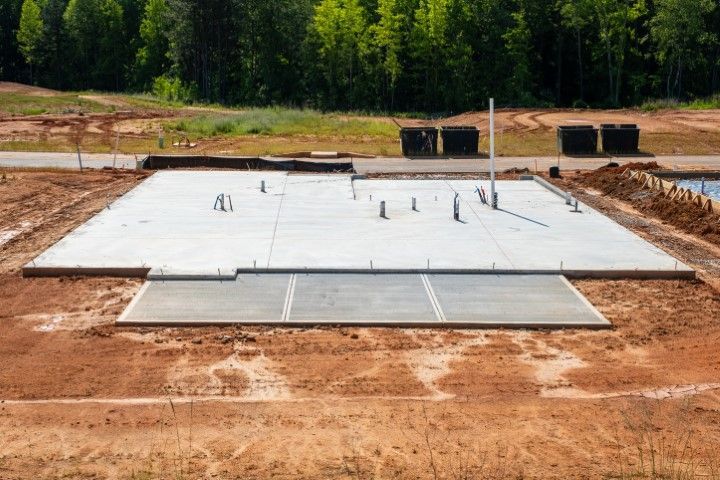 Concrete slab foundation of a house under construction in an open, dirt-covered lot with a treeline in the background.