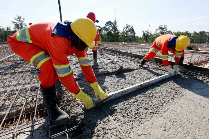 Construction workers in orange safety gear and yellow hard hats use a long level to smooth wet concrete on a metal frame.