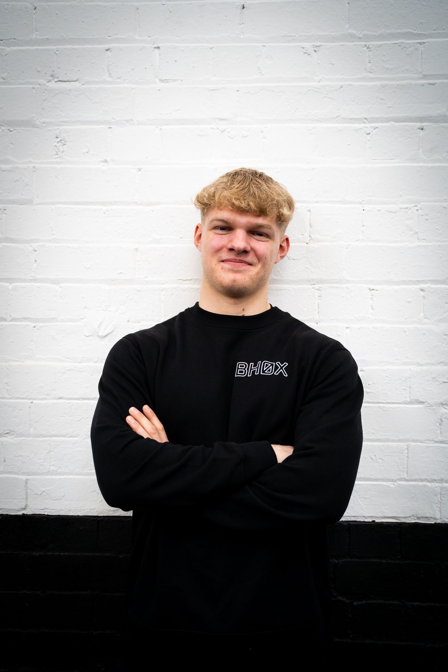 Blonde man in black long-sleeved shirt with arms crossed, smiling, standing in front of a white brick wall.