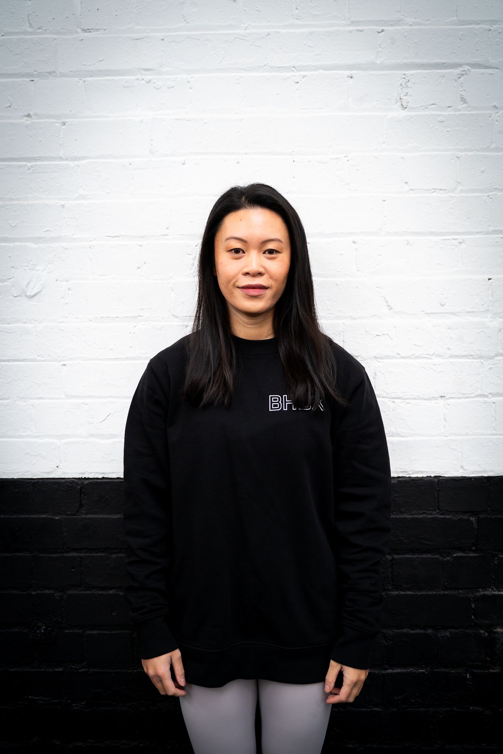 Woman standing in front of a black and white wall, wearing a black sweatshirt and light gray pants.