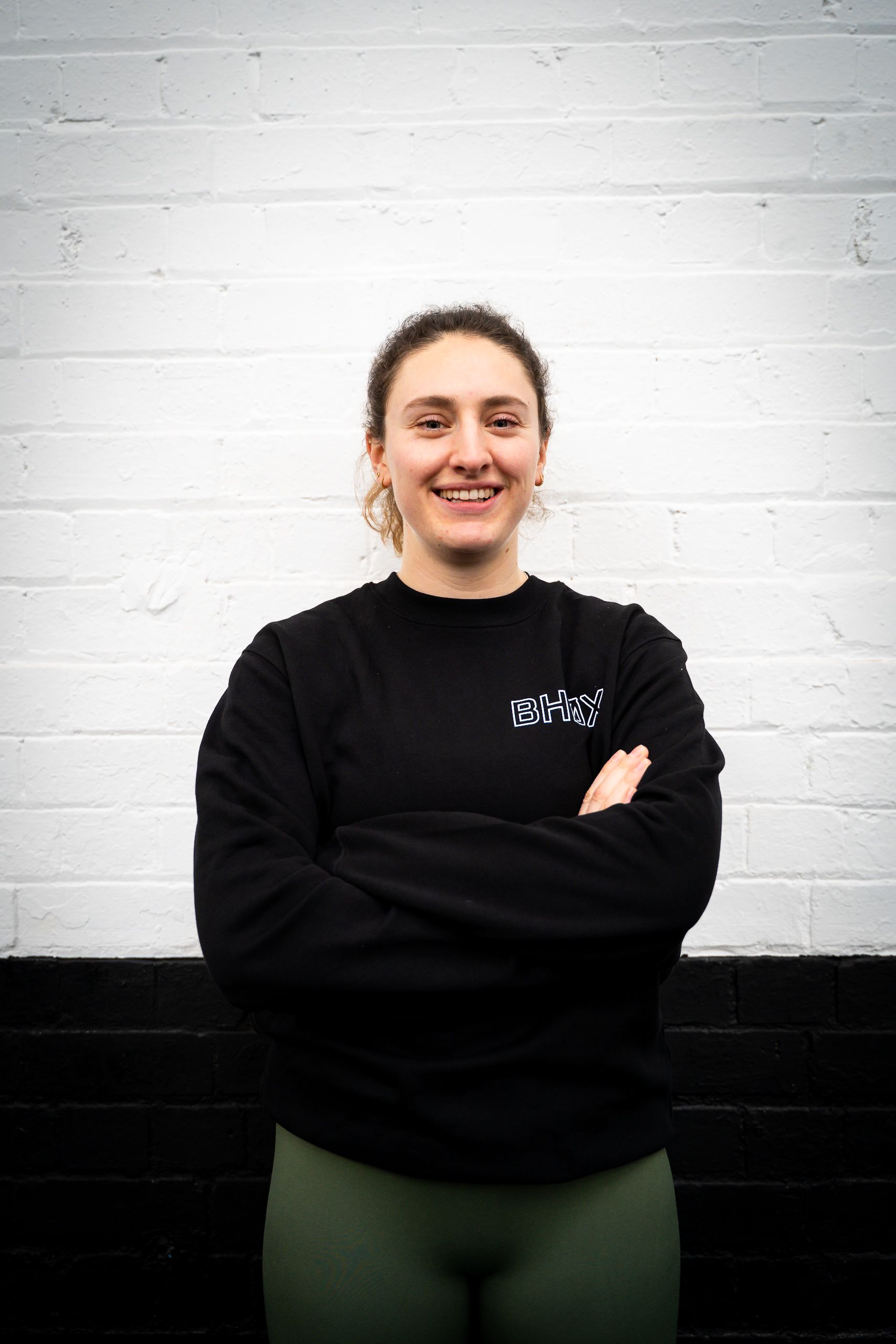 Woman in black sweatshirt and green leggings, arms crossed, smiling, in front of a white and black wall.
