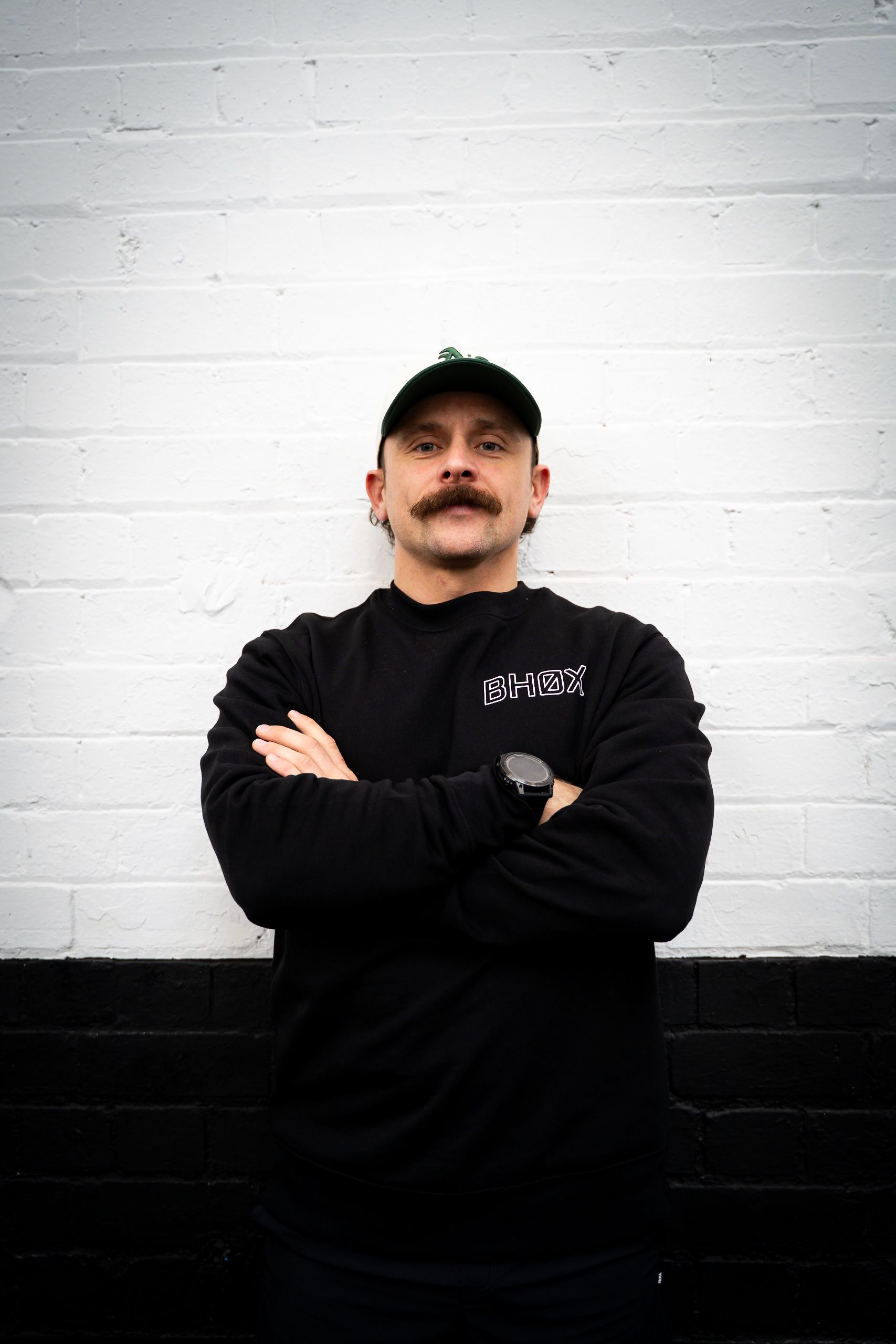 Man in black sweatshirt and cap, arms crossed, stands against a black and white brick wall.