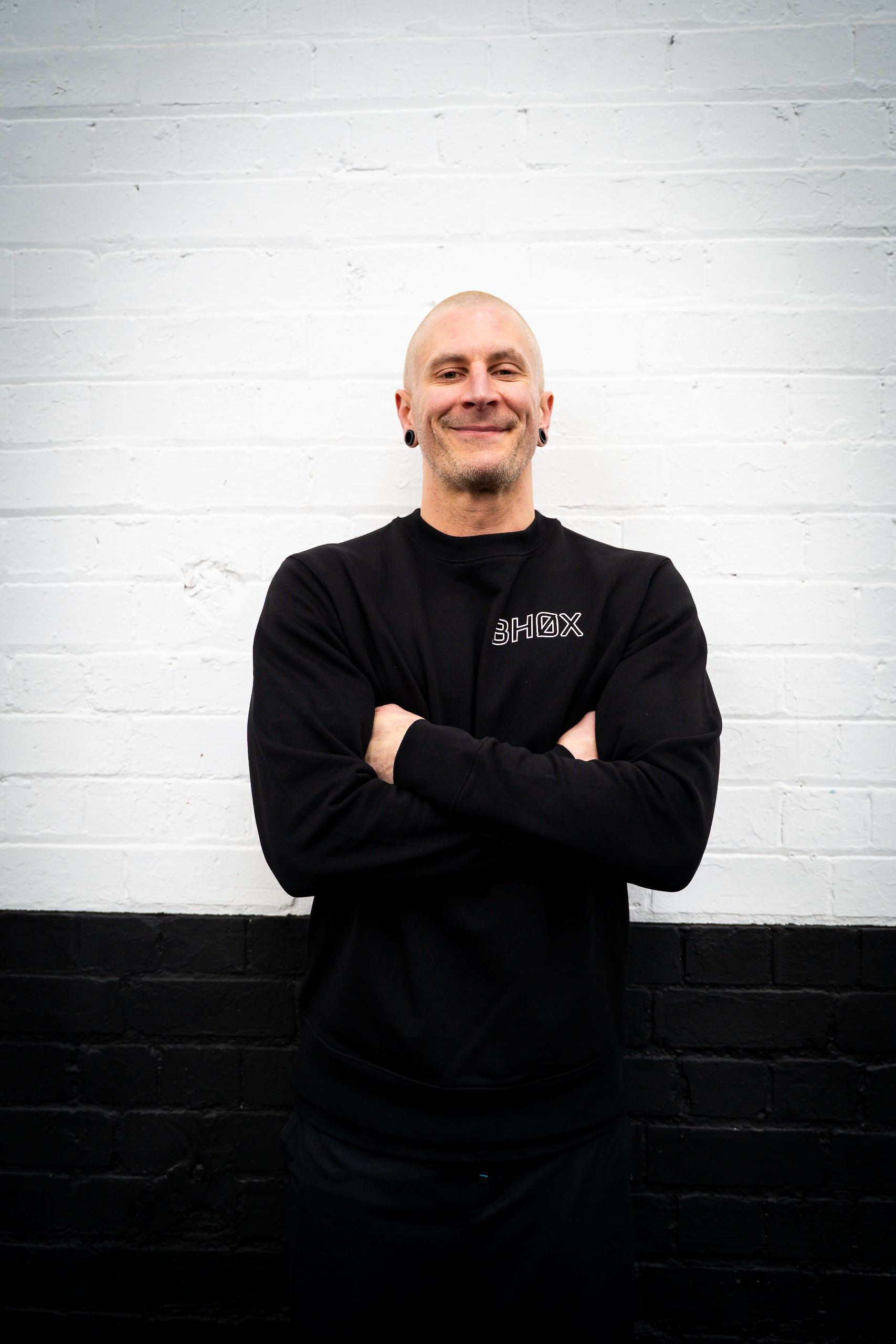 Man in black sweatshirt with arms crossed, smiling, in front of a white and black brick wall.