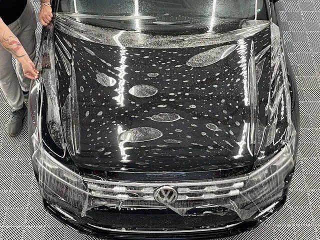 A man is covering the hood of a black car with plastic wrap.