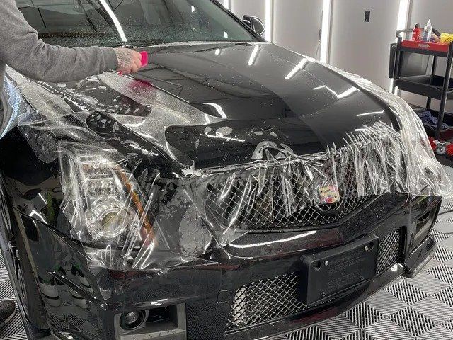 A man is covering the hood of a black car with plastic wrap.