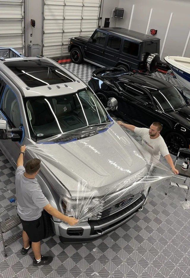 Two men are wrapping a silver suv with plastic wrap in a garage.