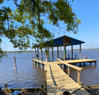 Wooden pier with a covered boat slip on a wide body of water, under a blue sky.