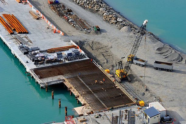 Construction site at a pier with workers, crane, and materials on the water.