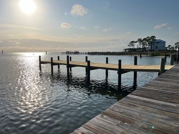 Wooden pier extends over calm water toward a sunny horizon; a building is visible in the distance.
