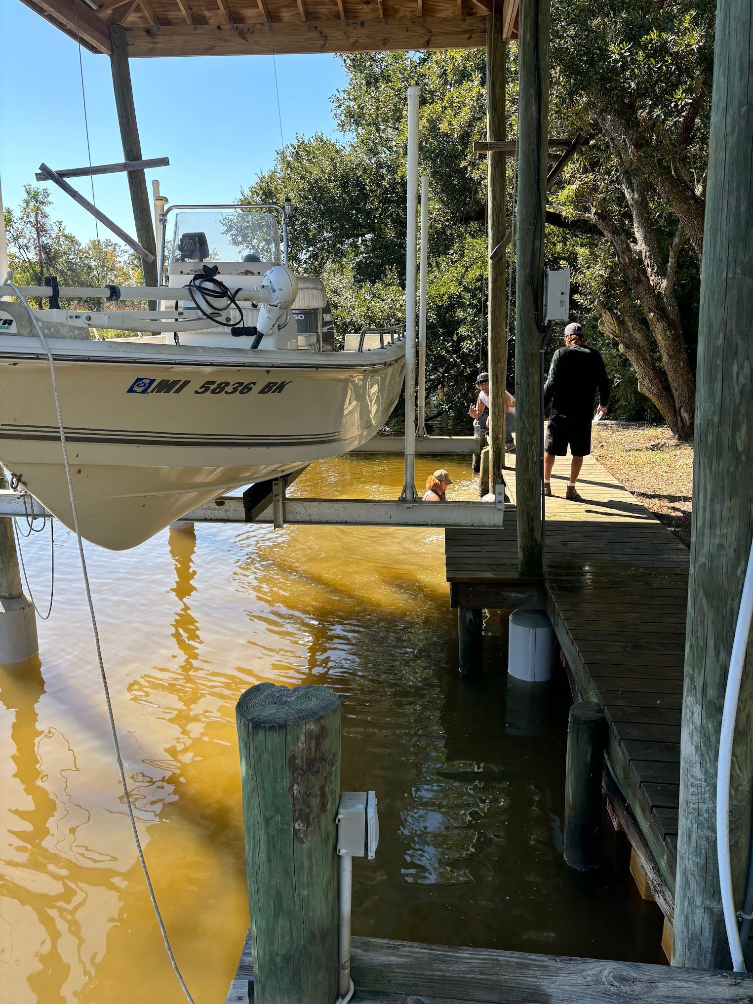 Boat on a lift over murky water at a dock, person walking on dock, sunny day.