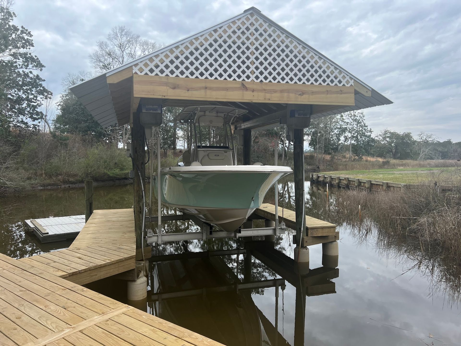 Boat on a lift in a covered boat dock. Dock has wooden deck and lattice roof. Water and trees in the background.