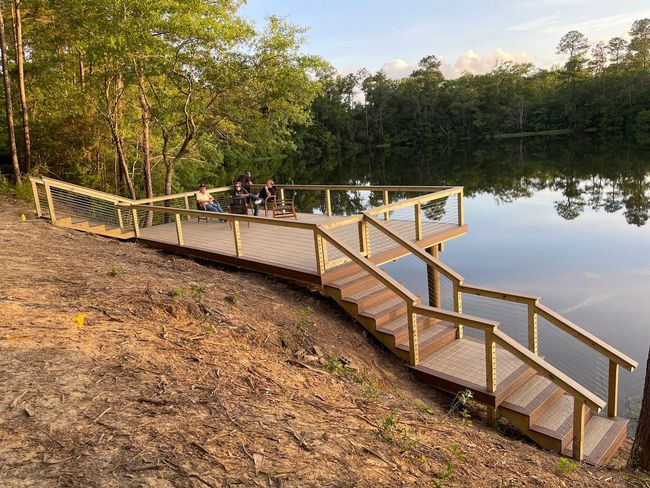 Wooden dock with steps leading to a calm lake, surrounded by trees. Two people sit on the dock.