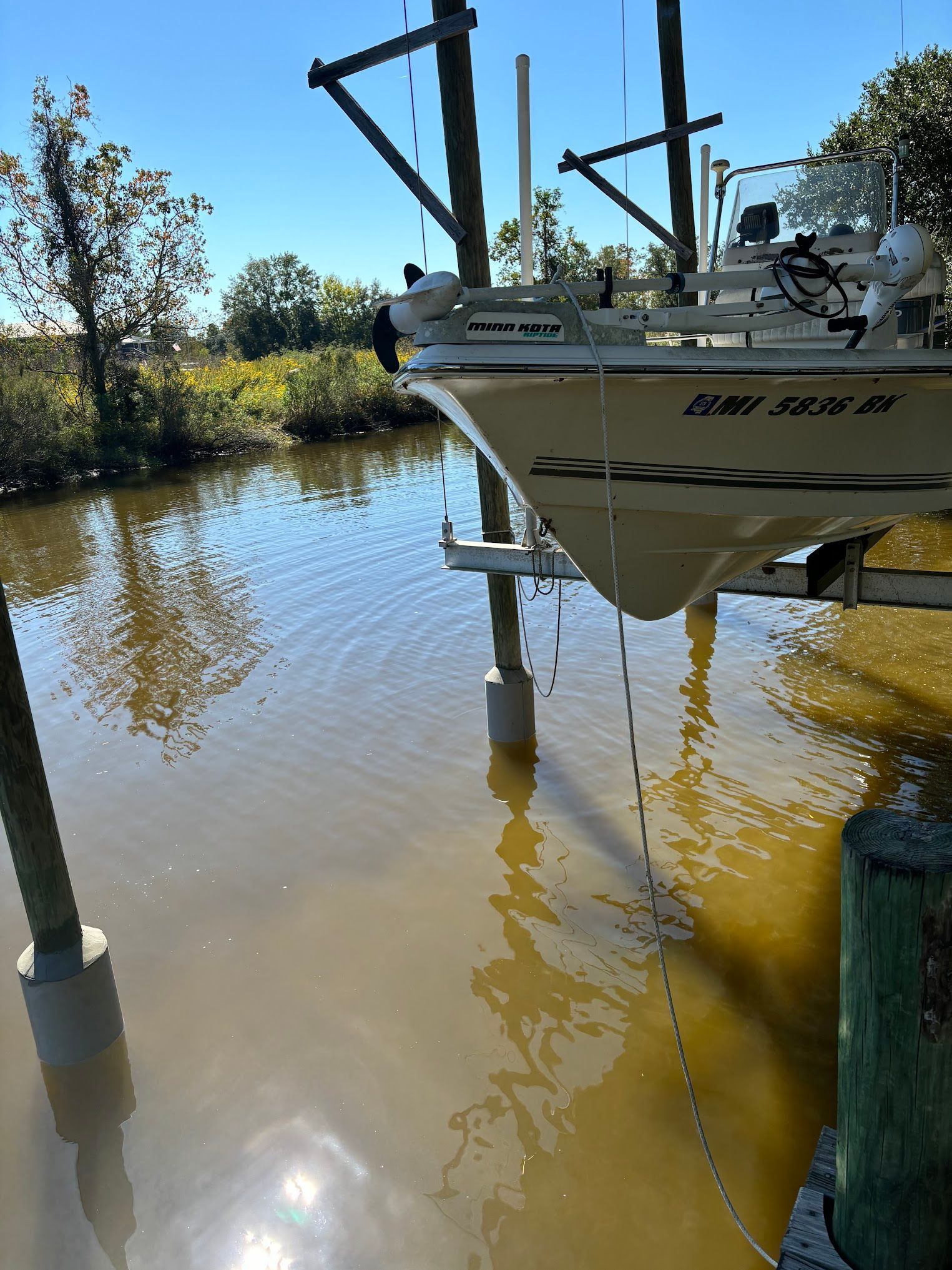 A boat on a lift over brown water, near green foliage under a sunny sky.