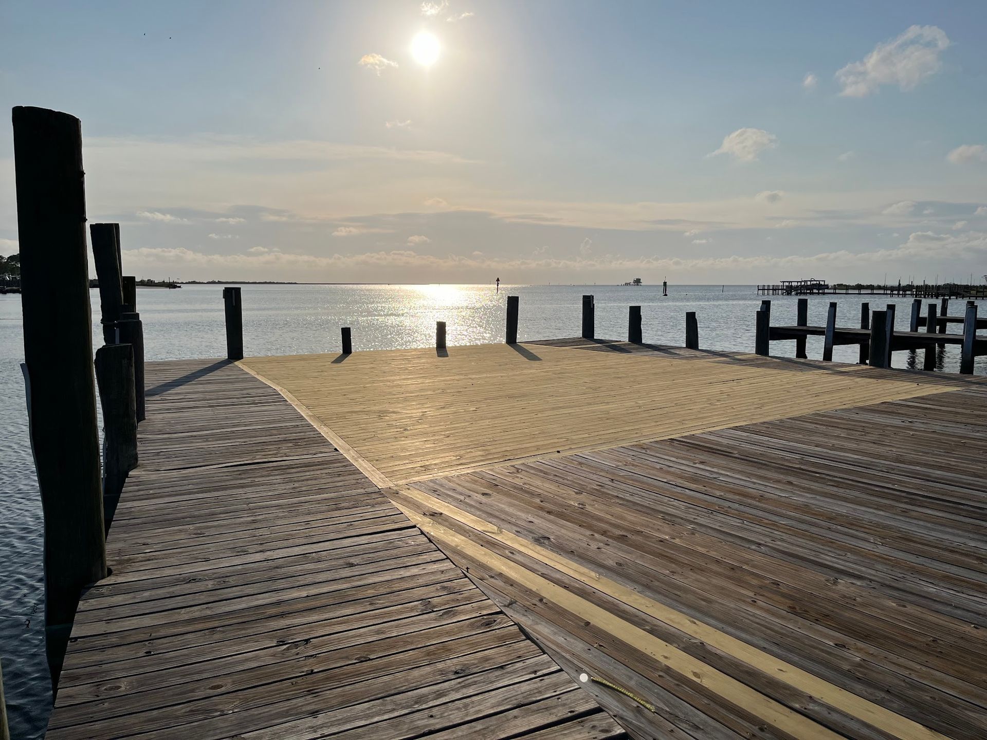 Wooden pier with sandy patch, leading to calm water under a bright sun.