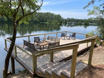 Wooden deck with seating overlooking a calm lake, surrounded by trees.
