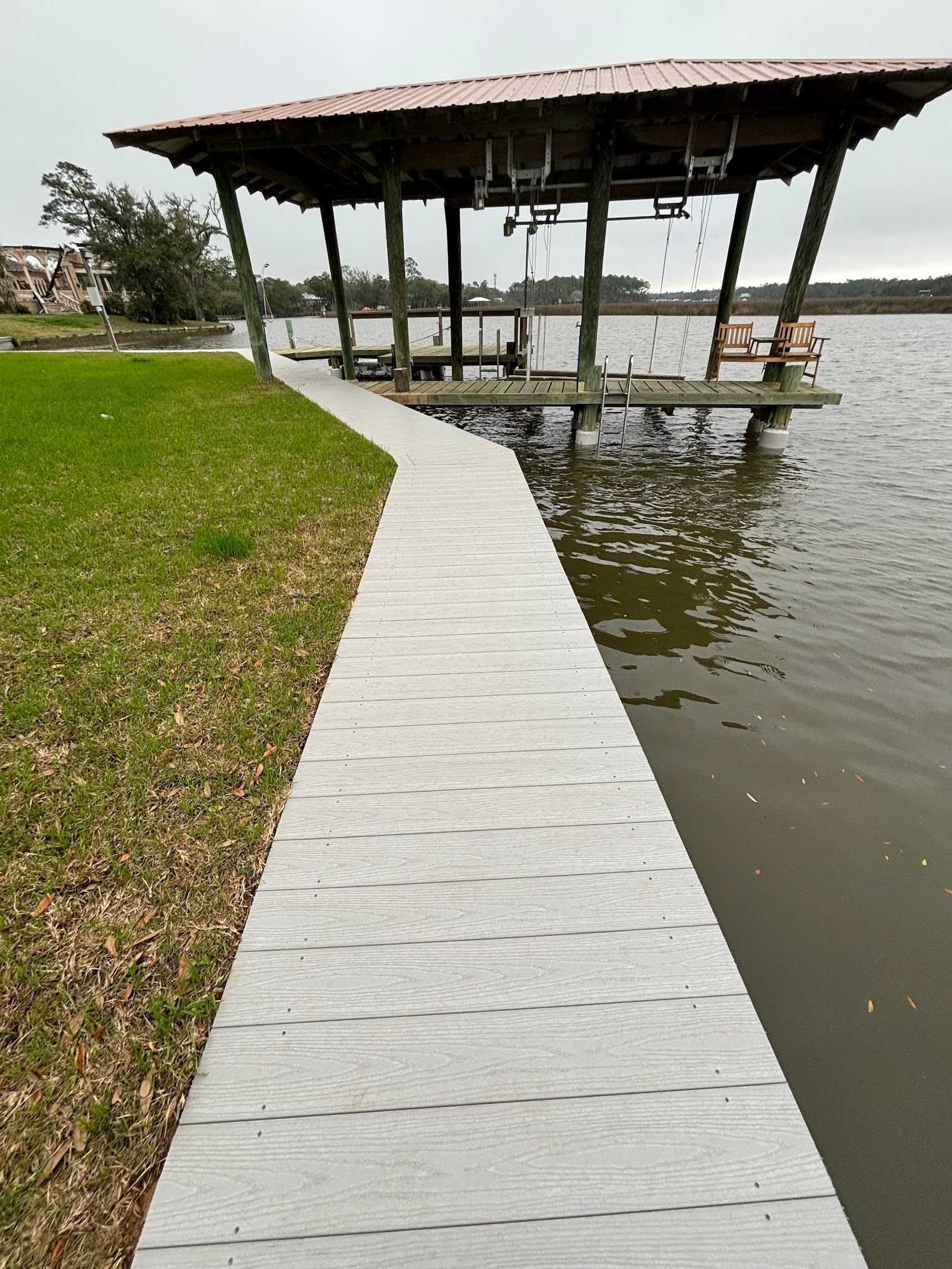 A gray walkway leads to a covered dock on a lake. Green grass borders the walkway.