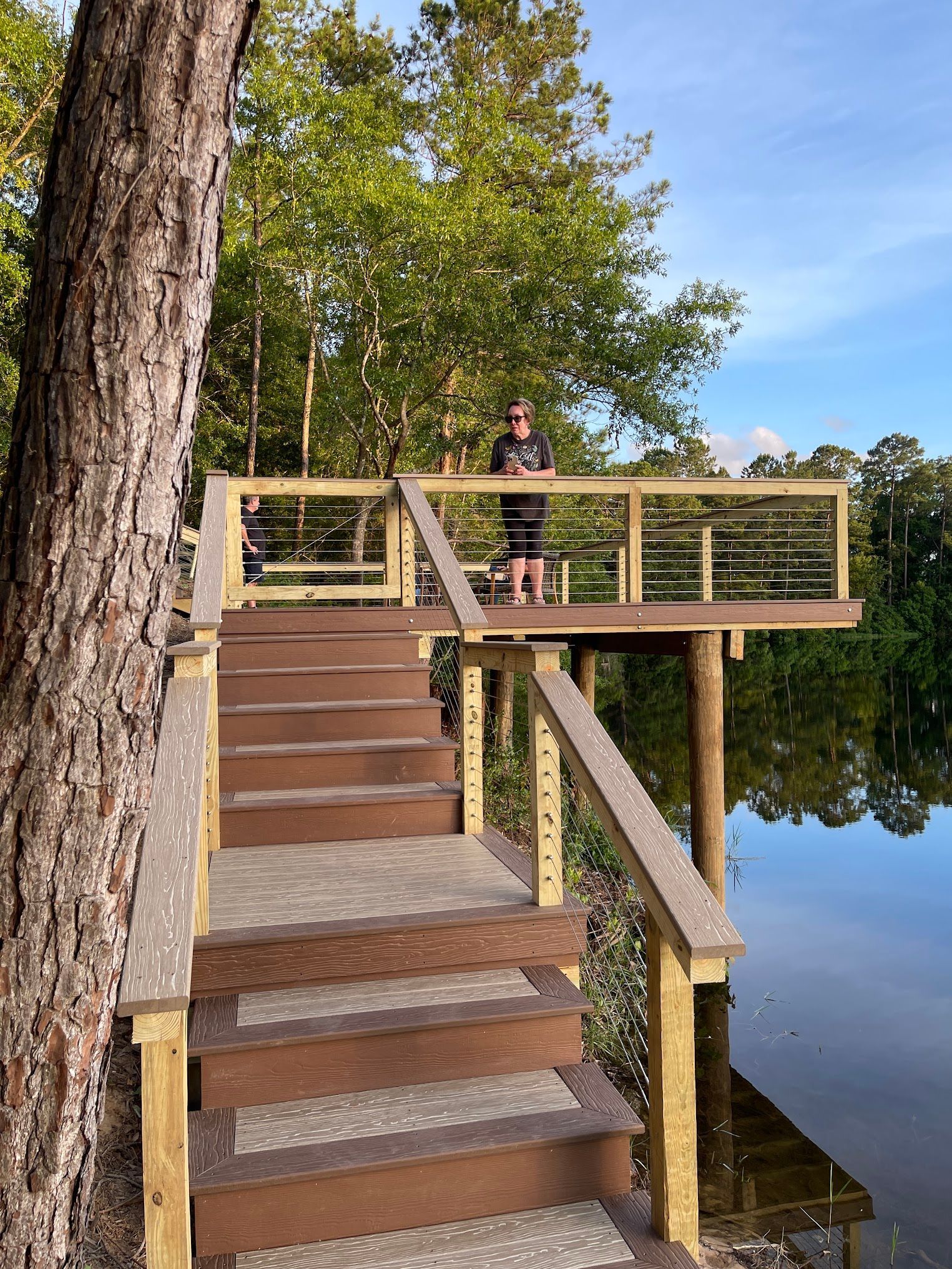 Person standing on a wooden platform over water; stairs lead up.  Trees and sky in the background.