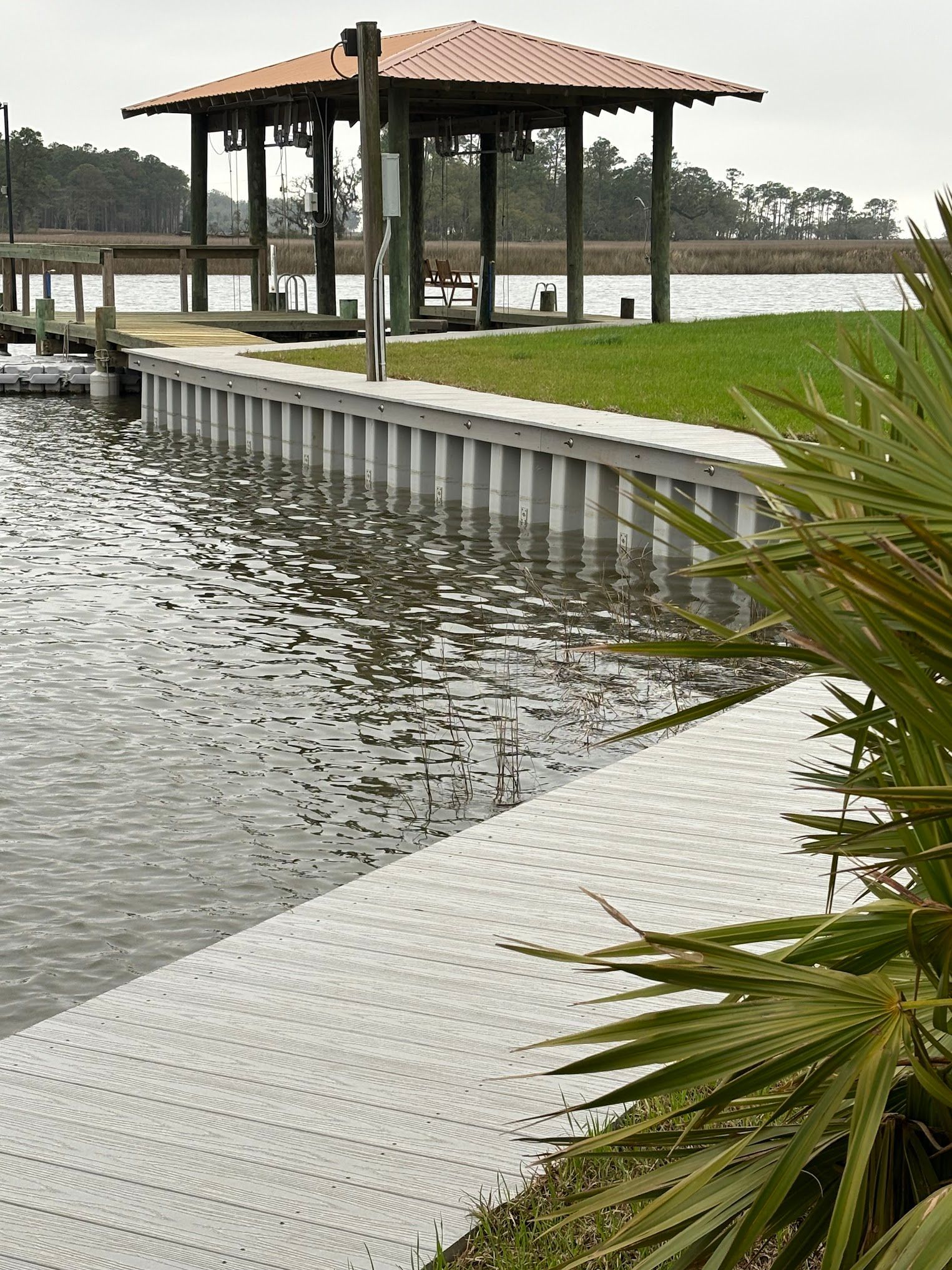 Dock and gazebo on a waterfront. Gray dock with gray retaining wall, brown roof, and green grass.