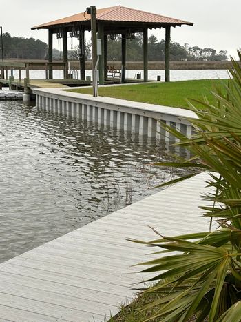 Dock and gazebo on a waterfront. Gray dock with gray retaining wall, brown roof, and green grass.