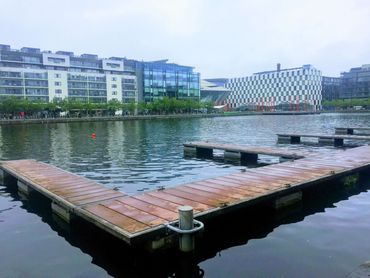 L-shaped wooden dock on water, modern buildings in the background under an overcast sky.