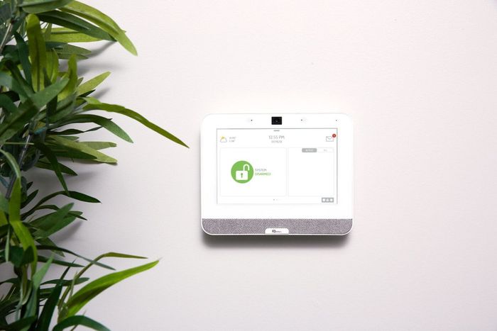 A smart device is sitting on a white wall next to a plant.