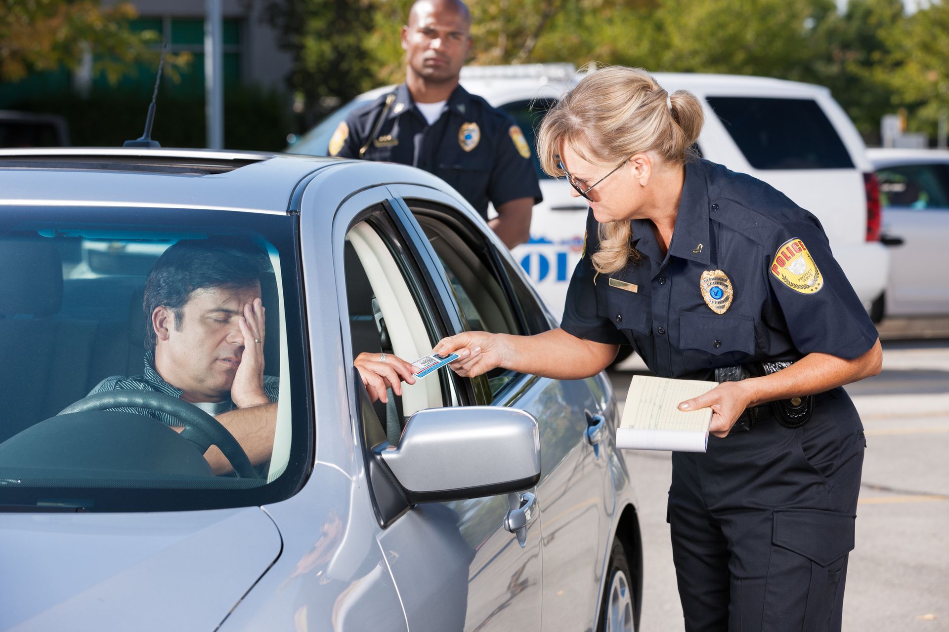 Police Officer Writing a Traffic Ticket - A mature female law enforcement officer stands by a vehicle she has stopped and takes the driver's license from a middle-aged man while her partner stands by in the background.