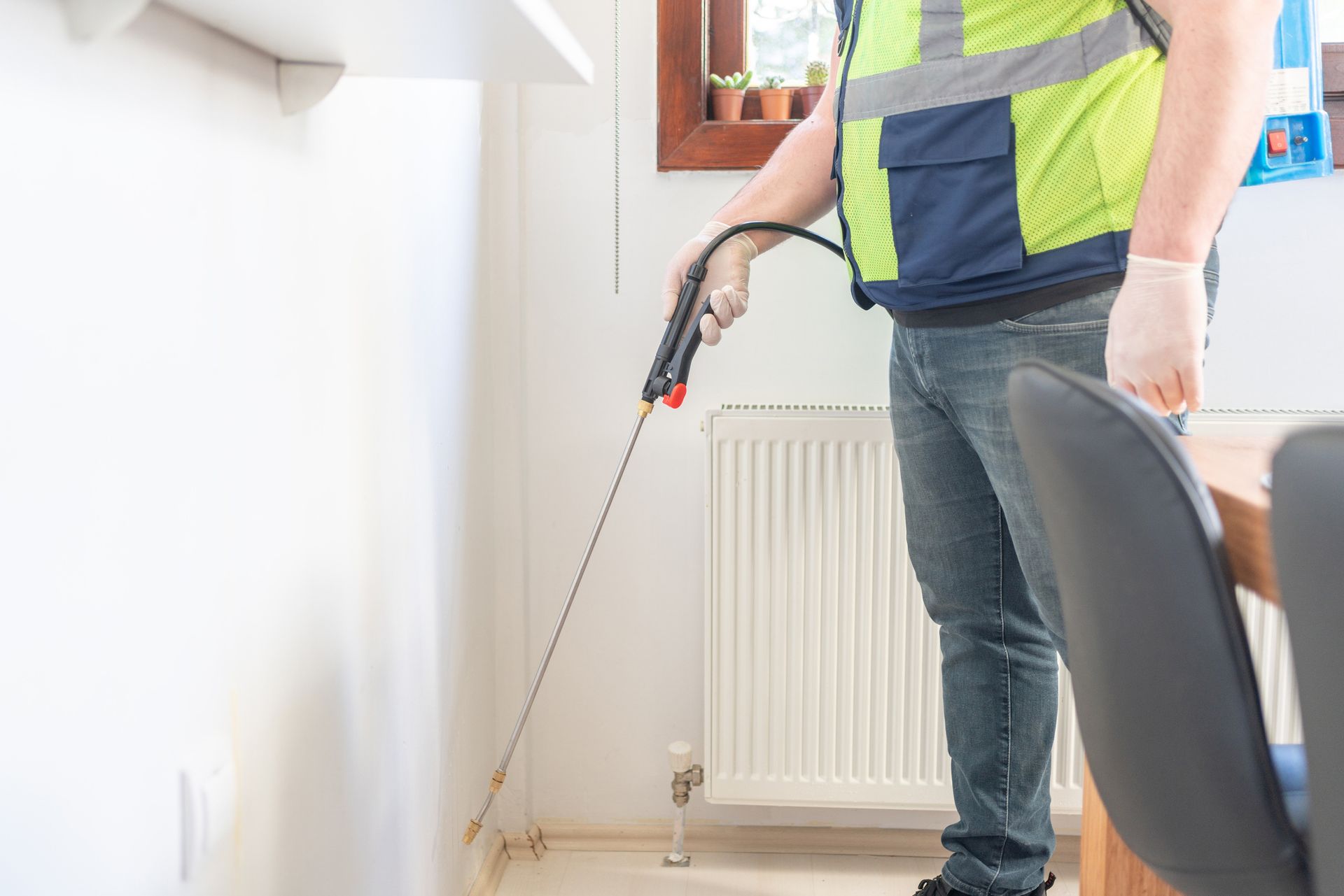 Person in safety vest spraying insecticide along a white wall indoors.