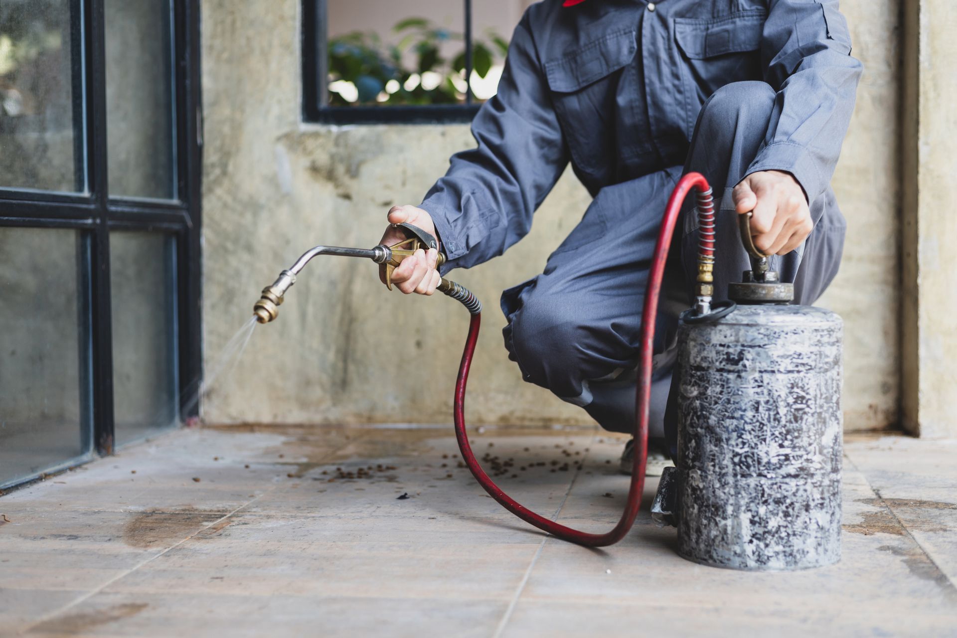 Pest control worker kneels, spraying insecticide from a tank. Gray overalls, outdoor setting, concrete floor.