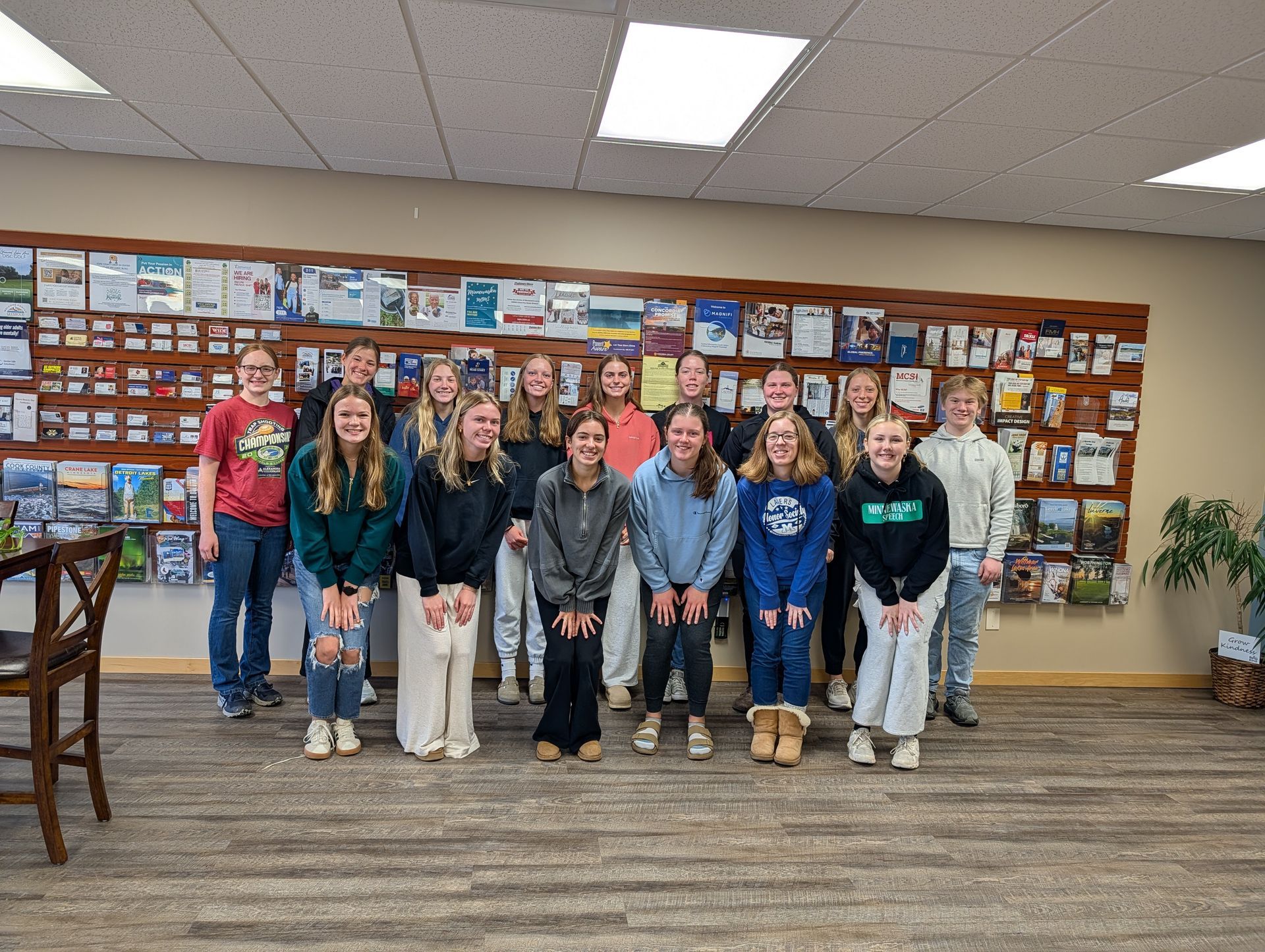 A group of 14 people stand in a line smiling in front of a wooden display rack filled with brochures in an office.